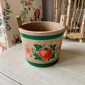 May include: A small, brown wooden bucket with green and pink floral designs. The bucket has two handles and is decorated with a large pink rose and smaller pink flowers.