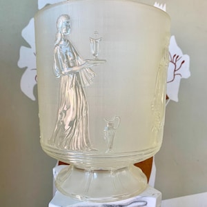 May include: A cylindrical frosted glass vase featuring a relief design of a figure in a long robe holding a tray with a small vessel. A pitcher is depicted near the figure's feet. The glass has a textured grid pattern and a flared base.