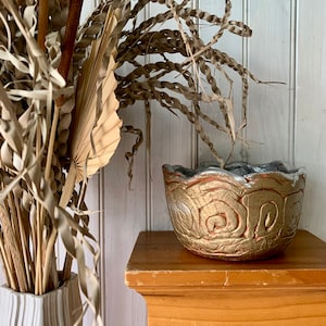 May include: A gold-painted ceramic planter with a wavy rim and a swirl design. The planter is sitting on a wooden shelf.
