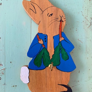 May include: Wooden bunny-shaped wall hook, painted with a blue jacket and holding carrots and greens. The bunny's ears and body are natural wood. A brass hook is attached for hanging items.