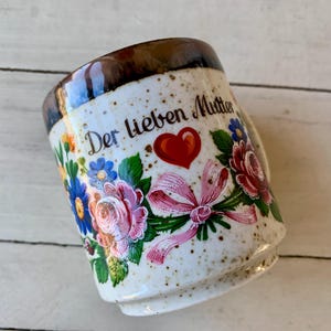 May include: A ceramic mug with a speckled cream finish and a dark brown rim. The front features a floral design with pink roses, blue flowers, a red heart, and a pink ribbon. The text "Der lieben Mutter" is printed in black script above the heart.