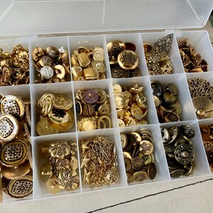 May include: A clear plastic organizer filled with various gold-toned buttons. The buttons vary in size, shape, and design, including floral, geometric, and ornate styles. Some buttons have a vintage aesthetic, while others appear more modern.