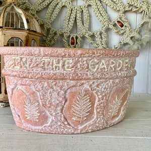 May include: A terracotta-colored planter with a raised rim and the words "In The Garden" embossed in gold lettering. The planter has a textured surface with fern leaf designs.