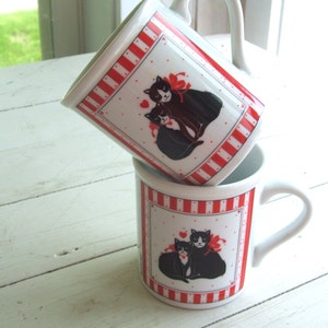 May include: Two white ceramic mugs with red and white stripes. Each mug features a black and white illustration of two cats with red bows. The cats are cuddling and have hearts around them.