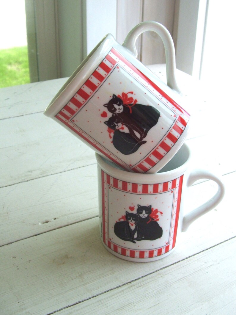 May include: Two white ceramic mugs with red and white stripes. Each mug features a black and white illustration of two cats with red bows. The cats are cuddling and have hearts around them.