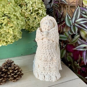 May include: A crocheted doll with a white dress and long hair. The doll is standing on a white surface with a pine cone and green plants in the background.