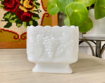 May include: White milk glass planter with a grapevine design. The planter has a square base and a scalloped rim.