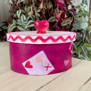 May include: A pink and white round box with a red heart-shaped knob on the lid. The box is decorated with a pink and white pattern of hearts and playing cards.