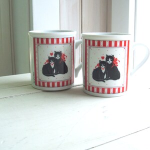 May include: Two white ceramic mugs with red and white striped bands around the top. Each mug features a black and white illustration of two cats with red bows, one cat is sitting on the other's back.