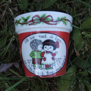 May include: A white ceramic Christmas mug with a red, green, and white design. It features a red ribbon with a bow and a cartoon image of two children. The text on the mug reads "All we need is love... it's Christmas time once more!"