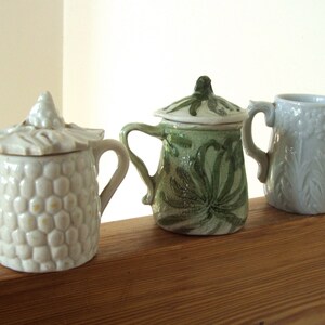 May include: Three ceramic mugs with different designs. The mug on the left is white with a textured surface. The mug in the middle is green with a leafy design. The mug on the right is light blue with a floral design.