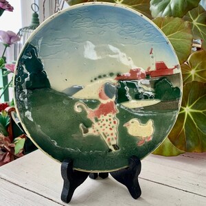 May include: A ceramic plate with a whimsical scene of a child with a duck. The plate features a green, blue, and cream color palette. The child is wearing a polka-dot outfit. The plate is displayed on a black wooden stand.