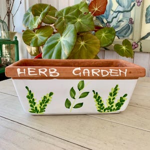 May include: Rectangular white ceramic herb garden planter with a brown rim. The words "HERB GARDEN" are painted in white on the rim. Green leaf and sprig designs are painted on the front. A green plant with large leaves is in the planter.