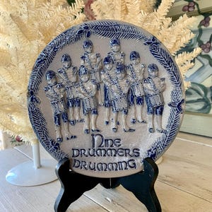 May include: A blue and white ceramic plate with a raised design of nine drummers playing drums. The plate has a blue and white border with a leafy design. The text "Nine Drummers Drumming" is written in blue on the plate.