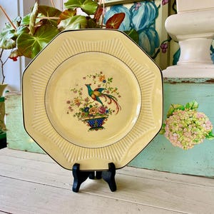 May include: Cream-colored octagonal plate with a black trim, featuring a central floral and bird design. The plate is displayed on a black stand, showcasing its decorative details. The plate's rim has a ribbed design.