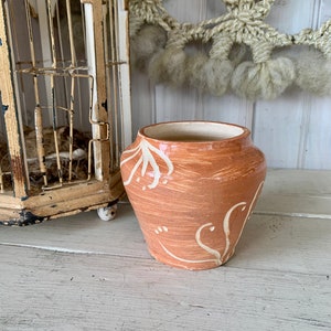 May include: A terracotta-colored ceramic vase with a white floral design. The vase has a rounded body and a slightly flared lip.