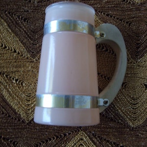 May include: A pink ceramic beer stein with a silver metal band around the top and bottom. The stein has a wooden handle.