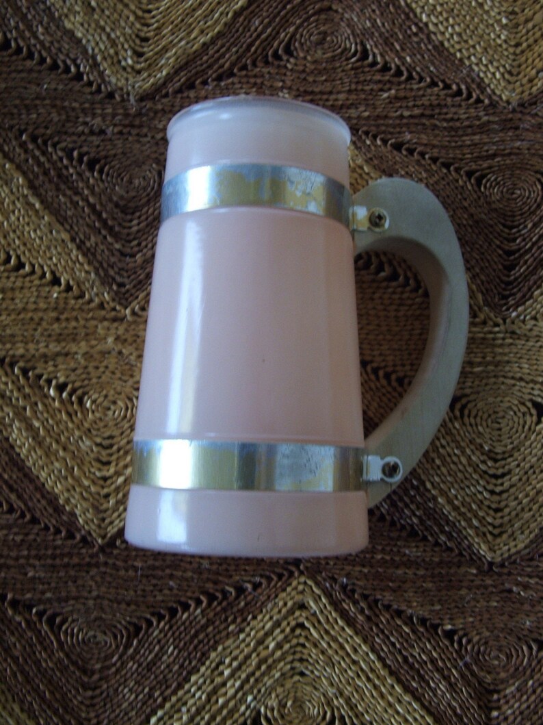 May include: A pink ceramic beer stein with a silver metal band around the top and bottom. The stein has a wooden handle.