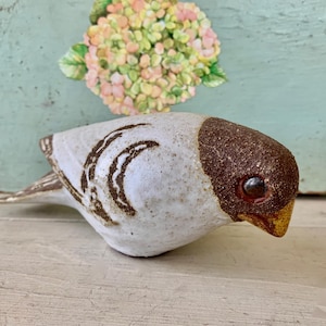May include: A ceramic bird figurine with a white body, a dark brown textured head, and a yellow beak. The wings feature brown painted markings, and the eye is a dark, glossy brown. The bird is positioned in profile, facing toward the right side.