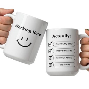 May include: Two white ceramic mugs. One mug has the text "Working Hard" with a smiling face. The other mug says "Actually:" with a checklist including "planning my dinner", "Internet shopping", "Booking a holiday", and "Job hunting".