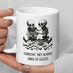 Pode incluir: Caneca de cerâmica branca com uma ilustração humorística de dois esqueletos sentados juntos, rodeados por ossos espalhados. Os esqueletos têm expressões tristes. O texto na caneca diz: "SOMEHOW, THEY ALWAYS ENDED UP LEGLESS".