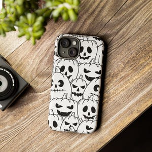 Puede incluir: Una funda de teléfono blanca con un patrón blanco y negro de caras de calabaza.