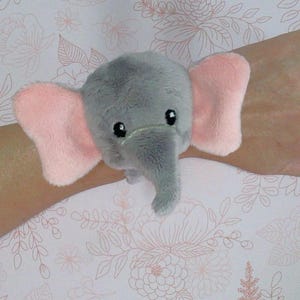 May include: A grey plush elephant with pink ears. The elephant is wearing a wristband and has a cute, cartoon-like face.