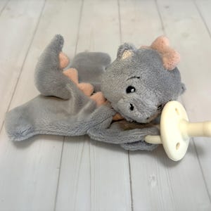 May include: A soft, plush grey dragon security blanket featuring a textured surface, light pink accents along the spine and head, and a cream-colored silicone pacifier attached to one corner of the fabric body with a round, flat shield.