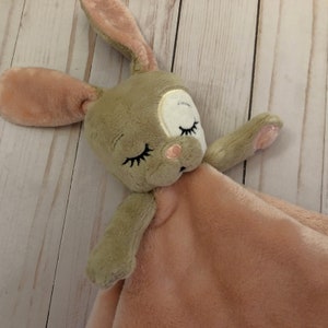 Puede incluir: Una manta de seguridad de peluche marrón y rosa con un conejo bordado con ojos y una nariz rosa. El conejo está acostado sobre una manta rosa.