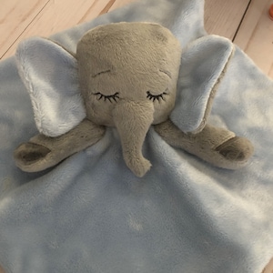 May include: A light blue baby blanket with a grey elephant sewn on top. The elephant has closed eyes and a small trunk.