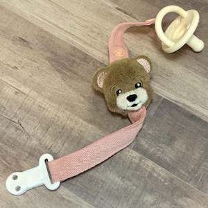 Op de afbeelding: Een roze en witte speenclip met een bruin teddybeerhoofd. De clip heeft een witte plastic clip en een roze stoffen band met een herringbone-patroon.