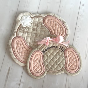 May include: Two quilted bunny-shaped pot holders. The larger one has a floral pattern and a white flower applique. The smaller one has the embroidered text "Bottoms Up" and a pink bow. Both have pink ear accents.