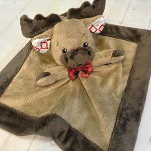 May include: A plush moose-shaped baby blanket in shades of brown and tan. The moose has a red and black plaid bow tie and patterned accents on its ears. The blanket has a soft, plush texture and a dark brown border.