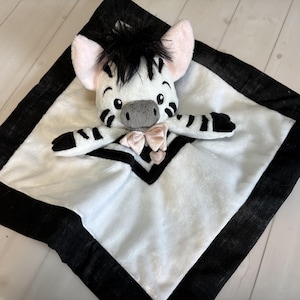 Pode incluir: Um cobertor de bebê com tema de zebra em preto e branco com uma cabeça e braços de zebra de pelúcia anexados. A zebra tem uma crina preta, orelhas rosa e um focinho cinza. Uma fita rosa está no pescoço da zebra. O cobertor tem uma borda preta.