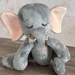 May include: A gray plush elephant toy with pink ears and closed eyes. The elephant is wearing a floral patterned bow tie and has a pink heart sewn on its chest.
