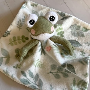 May include: A plush green frog toy with large white eyes and a pink heart on a soft, white blanket with green leaf patterns. The frog has a white belly and pink cheeks. The blanket is a square shape.
