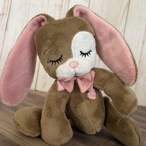Puede incluir: Un conejito de peluche marrón con orejas rosas y un lazo rosa. El conejito tiene pelaje blanco alrededor de los ojos y una nariz rosa. El conejito está sentado con los ojos cerrados.