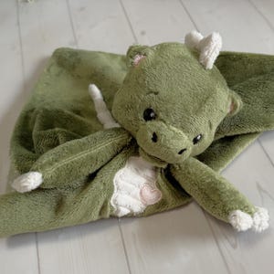 May include: A plush security blanket featuring a soft, olive green dragon head with embroidered black eyes and nostrils. The blanket body is made of matching olive green plush fabric with white textured accents on the dragon's back and a small, light pink heart patch on the front center.
