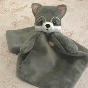 May include: A gray plush security blanket with a fox head attached. The fox has a white face with black eyes and a pink nose. The blanket has a pink heart on the front.