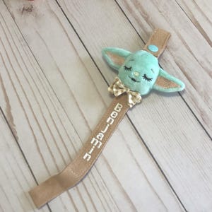 May include: A baby pacifier clip featuring a plush, light teal character head with closed eyes and a bow tie. The tan strap has the name "Benjamin" embroidered in white. A light blue button secures the clip.