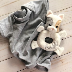 May include: A gray baby onesie with a white and brown puppy rattle toy. The rattle is shaped like a donut and has the word "Puppy" embroidered on it.