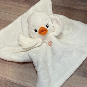 Può includere: Coperta per neonati in peluche bianca con un'anatra bianca attaccata. L'anatra ha un becco arancione e occhi neri. Un cuore rosa è ricamato sulla coperta.