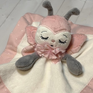 Op de afbeelding: Een zachte vlinderknuffel in roze, wit en grijs. De vlinder heeft gesloten ogen, een geplooid kraagje en grijze antennes en armen. De deken heeft een roze rand met witte stippen.
