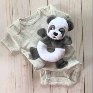 Pode incluir: Um macacão de bebê cinza e branco com um chocalho de urso panda. O chocalho é branco com um anel preto e um rosto de urso panda.