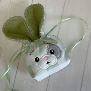 Puede incluir: Una pequeña bolsa con cordón de ajuste con temática de conejito, a cuadros verdes claros y blancos. La bolsa presenta una cara de conejito con ojos negros, una nariz rosa y un parche gris en la oreja. La cinta verde se utiliza como cordón y las orejas de conejo son verdes.