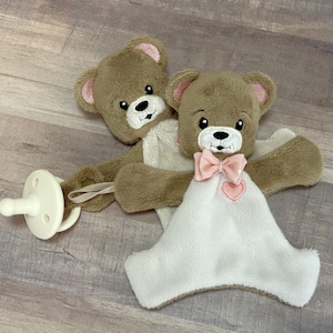 Op de afbeelding: Een bruine teddybeer knuffel met een witte deken, een roze strik en een fopspeen. De beer heeft roze oren en een hartvormige patch. De fopspeen is wit.