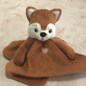 May include: A brown plush fox security blanket with a pink heart on the front. The fox has a white face with black eyes and a black nose.