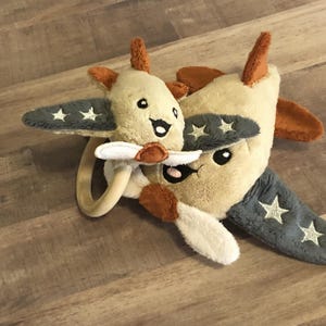 Puede incluir: Dos juguetes de avión de peluche, uno más grande que el otro, con alas grises y estrellas blancas. El avión más pequeño tiene un anillo de madera unido a él. Ambos aviones tienen caras sonrientes.