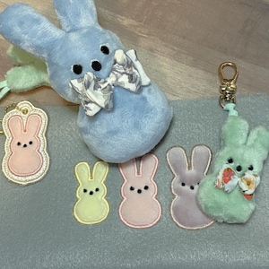 Puede incluir: Un conejito de peluche azul claro con una pajarita blanca y floral, rodeado de cinco formas de conejito bordadas en colores pastel sobre un fondo de tela azul claro.