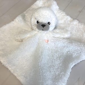 May include: A white plush baby security blanket with a seal head. The seal has gray accents on its face and a black nose. The blanket is soft and fluffy, with a pink heart-shaped detail.
